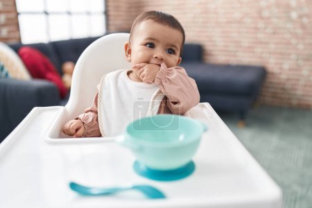 Foto de Adorable bebé hispano chupando el dedo sentado en la silla alta en casa - Imagen libre de derechos