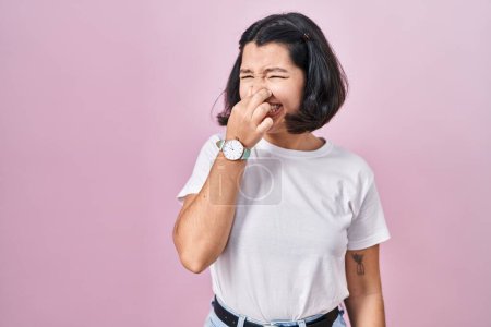 Foto de Young hispanic woman wearing casual white t shirt over pink background smelling something stinky and disgusting, intolerable smell, holding breath with fingers on nose. bad smell - Imagen libre de derechos