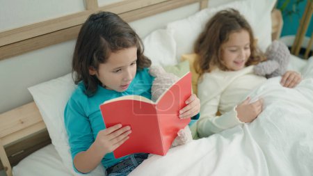 Foto de Two kids reading story book sitting on bed at bedroom - Imagen libre de derechos