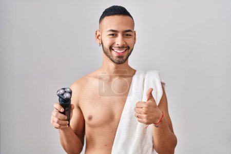 Téléchargez les photos : Jeune homme hispanique tenant rasoir électrique souriant heureux et positif, pouce levé faire excellent et signe d'approbation - en image libre de droit