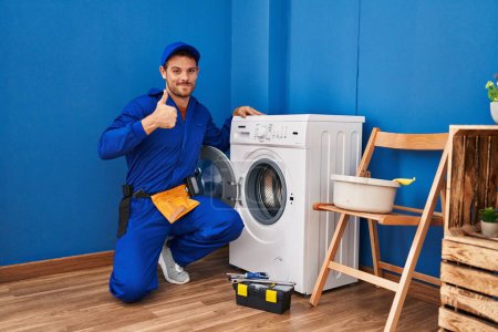 Photo for Young hispanic man working on washing machine smiling happy and positive, thumb up doing excellent and approval sign - Royalty Free Image