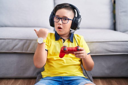 Photo for Young hispanic kid playing video game holding controller wearing headphones surprised pointing with hand finger to the side, open mouth amazed expression. - Royalty Free Image