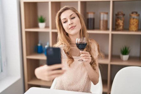 Foto de Joven mujer rubia bebiendo vino haciendo selfie junto al smartphone en casa - Imagen libre de derechos