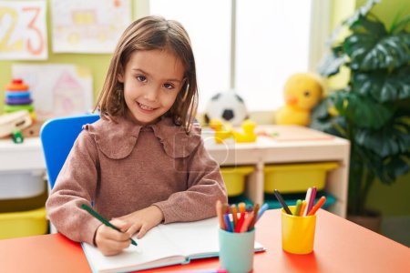Foto de Adorable hispanic girl preschool student sitting on table writing on notebook at kindergarten - Imagen libre de derechos