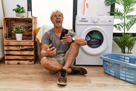 Téléchargez les photos : Jeune homme blond faisant la lessive en utilisant smartphone en colère et fou hurlant frustré et furieux, criant de colère. rage et concept agressif. - en image libre de droit