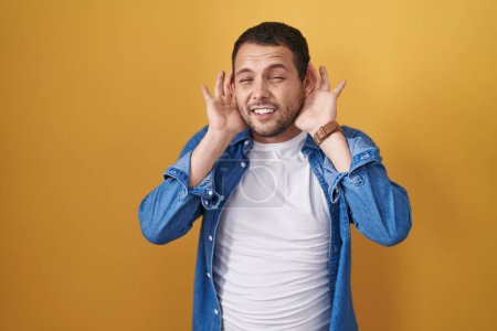 Photo for Hispanic man standing over yellow background trying to hear both hands on ear gesture, curious for gossip. hearing problem, deaf - Royalty Free Image