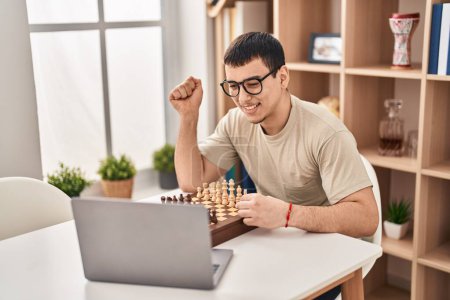 Foto de Hombre árabe joven jugando ajedrez en línea gritando orgulloso, celebrando la victoria y el éxito muy emocionado con el brazo levantado - Imagen libre de derechos