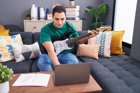 Téléchargez les photos : Jeune homme hispanique ayant cours de guitare électrique en ligne assis sur le canapé à la maison - en image libre de droit