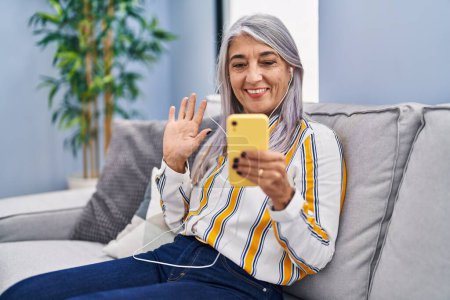 Téléchargez les photos : Femme d'âge moyen avec les cheveux gris en utilisant smartphone assis sur le canapé regardant positif et heureux debout et souriant avec un sourire confiant montrant les dents - en image libre de droit