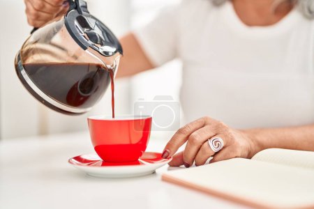 Téléchargez les photos : Femme d'âge moyen versant du café sur la tasse assise sur la table à la maison - en image libre de droit