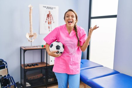Téléchargez les photos : Young hispanic woman working at physiotherapy clinic holding football ball celebrating victory with happy smile and winner expression with raised hands - en image libre de droit