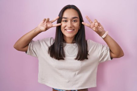 Téléchargez les photos : Jeune femme hispanique debout sur fond rose faisant symbole de paix avec les doigts sur le visage, souriant joyeuse montrant la victoire - en image libre de droit