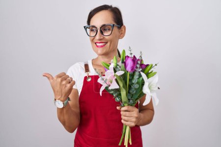 Foto de Mujer morena de mediana edad vistiendo delantal trabajando en floristería sosteniendo ramo sonriendo con cara feliz mirando y señalando a un lado con el pulgar hacia arriba. - Imagen libre de derechos