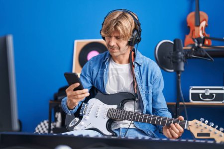 Téléchargez les photos : Jeune homme artiste utilisant smartphone tenant guitare électrique au studio de musique - en image libre de droit