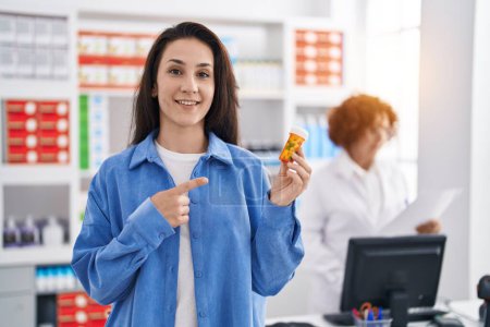 Téléchargez les photos : Young brunette woman holding pills at the pharmacy smiling happy pointing with hand and finger - en image libre de droit