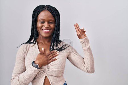 Foto de Mujer africana con trenzas de pie sobre fondo blanco sonriendo jurando con la mano en el pecho y los dedos hacia arriba, haciendo una promesa de lealtad juramento - Imagen libre de derechos