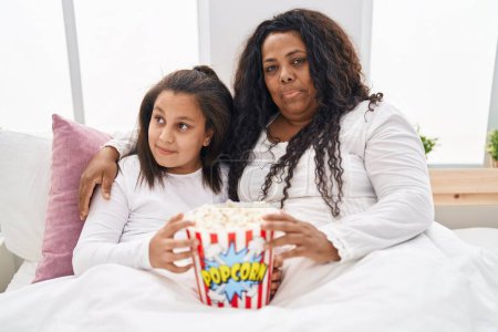 Téléchargez les photos : Mère et jeune fille mangeant du pop-corn dans le lit souriant regardant vers le côté et regardant loin en pensant. - en image libre de droit