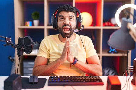 Photo for Hispanic man with beard playing video games with headphones begging and praying with hands together with hope expression on face very emotional and worried. begging. - Royalty Free Image