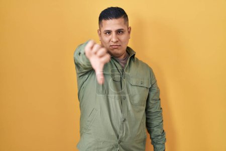 Foto de Hispanic young man standing over yellow background looking unhappy and angry showing rejection and negative with thumbs down gesture. bad expression. - Imagen libre de derechos