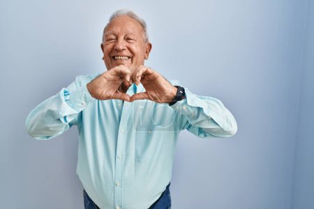 Photo for Senior man with grey hair standing over blue background smiling in love doing heart symbol shape with hands. romantic concept. - Royalty Free Image