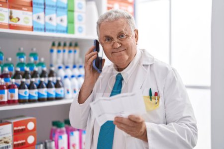 Photo for Middle age grey-haired man pharmacist talking on smartphone reading prescription at pharmacy - Royalty Free Image