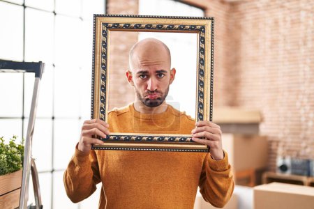 Foto de Young bald man with beard moving to a new home putting face inside vintage frame depressed and worry for distress, crying angry and afraid. sad expression. - Imagen libre de derechos