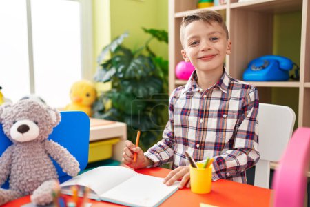 Foto de Adorable estudiante chico caucásico sentado en el dibujo de mesa en el cuaderno en el jardín de infantes - Imagen libre de derechos