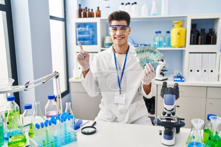 Photo for Young hispanic man working at scientist laboratory holding brazilian reals smiling happy pointing with hand and finger to the side - Royalty Free Image