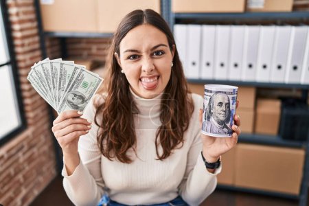 Téléchargez les photos : Young hispanic woman working at small business ecommerce holding money and piggy bank sticking tongue out happy with funny expression. - en image libre de droit