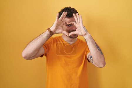 Photo for Young hispanic man standing over yellow background doing heart shape with hand and fingers smiling looking through sign - Royalty Free Image