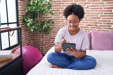 Téléchargez les photos : Femme afro-américaine utilisant pavé tactile assis sur le lit dans la chambre - en image libre de droit