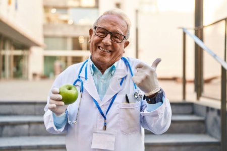 Foto de Médico senior con cabello gris sosteniendo saludable manzana verde sonriendo feliz y positivo, pulgar hacia arriba haciendo excelente y signo de aprobación - Imagen libre de derechos