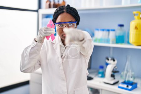 Foto de Mujer afroamericana trabajando en laboratorio científico sosteniendo cinta rosa señalando con el dedo a la cámara y a ti, gesto confiado que parece serio - Imagen libre de derechos