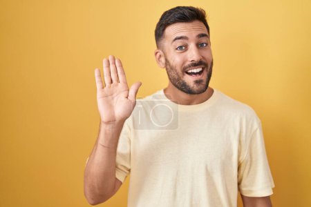 Téléchargez les photos : Bel homme hispanique debout sur fond jaune renonçant à dire bonjour heureux et souriant, geste de bienvenue amical - en image libre de droit