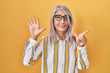 Téléchargez les photos : Femme d'âge moyen avec les cheveux gris debout sur fond jaune portant des lunettes montrant et pointant vers le haut avec les doigts numéro sept tout en souriant confiant et heureux. - en image libre de droit