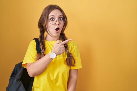 Téléchargez les photos : Young caucasian woman wearing student backpack over yellow background surprised pointing with finger to the side, open mouth amazed expression. - en image libre de droit