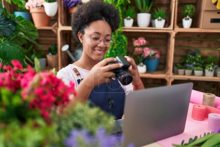 Photo for African american woman florist using laptop holding professional camera at flower shop - Royalty Free Image