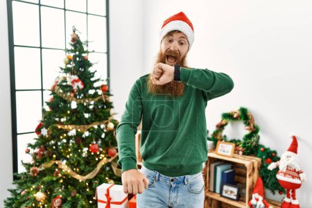 Foto de Pelirrojo con barba larga usando sombrero de navidad junto al árbol de navidad mirando el tiempo del reloj preocupado, con miedo de llegar tarde - Imagen libre de derechos