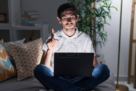 Photo for Young hispanic man using laptop at home at night pointing up looking sad and upset, indicating direction with fingers, unhappy and depressed. - Royalty Free Image