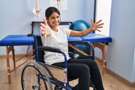 Photo for Young hispanic woman sitting on wheelchair at physiotherapy clinic looking at the camera smiling with open arms for hug. cheerful expression embracing happiness. - Royalty Free Image