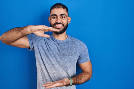 Téléchargez les photos : Middle east man with beard standing over blue background gesturing with hands showing big and large size sign, measure symbol. smiling looking at the camera. measuring concept. - en image libre de droit