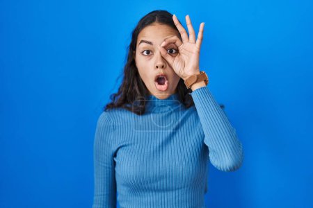 Foto de Young brazilian woman standing over blue isolated background doing ok gesture shocked with surprised face, eye looking through fingers. unbelieving expression. - Imagen libre de derechos