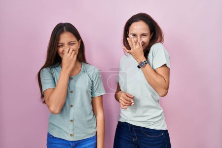 Téléchargez les photos : Jeune mère et fille debout sur fond rose sentant quelque chose de puant et dégoûtant, odeur intolérable, retenant la respiration avec les doigts sur le nez. mauvaise odeur - en image libre de droit