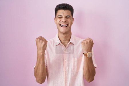 Foto de Joven hombre hispano de pie sobre fondo rosa emocionado por el éxito con los brazos levantados y los ojos cerrados celebrando la victoria sonriendo. concepto ganador. - Imagen libre de derechos
