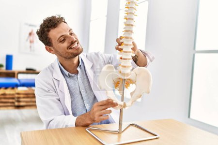 Photo for Young hispanic man wearing physiotherapist uniform touching anatomical model of vertebral column at clinic - Royalty Free Image