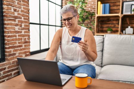 Téléchargez les photos : Femme d'âge moyen utilisant un ordinateur portable et une carte de crédit à la maison - en image libre de droit