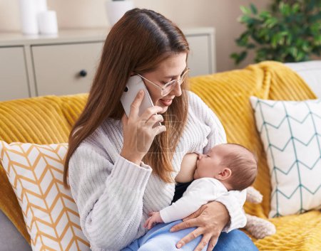 Photo for Mother and son talking on smartphone breastfeeding baby at home - Royalty Free Image