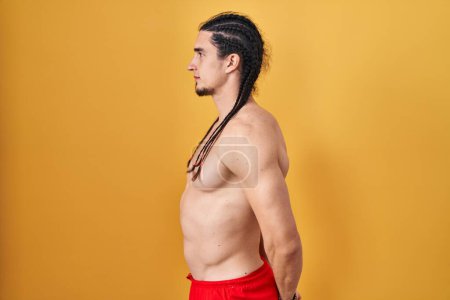 Foto de Hispanic man with long hair standing shirtless over yellow background looking to side, relax profile pose with natural face and confident smile. - Imagen libre de derechos