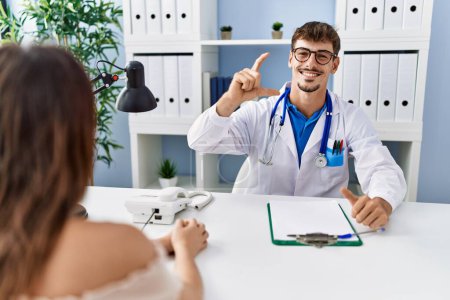 Foto de Young doctor with client at medical clinic smiling and confident gesturing with hand doing small size sign with fingers looking and the camera. measure concept. - Imagen libre de derechos
