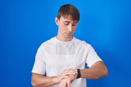 Foto de Hombre rubio caucásico de pie sobre fondo azul comprobando la hora en el reloj de pulsera, relajado y seguro - Imagen libre de derechos